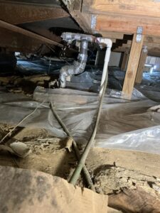 Plumbing beneath a mobile home, with exposed wood framing, wrapped pipes, and plastic ground covering.